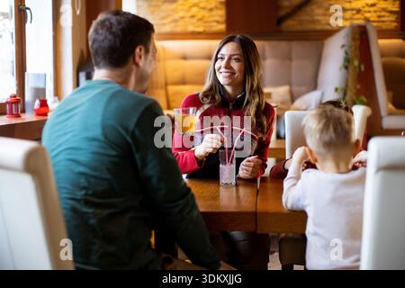 Vier Personen sitzen an einem Holztisch in einem Café, genießen Drinks und plaudern. Zwei Erwachsene und zwei Kinder unterhalten sich mit einem Lächeln Stockfoto