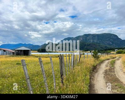 Malerische Sommerlandschaft mit ländlicher Feldstraße, die entlang eines alten hölzernen Viehzaun und einer breiten Wiese mit wilden gelben Blumen verläuft. Stockfoto