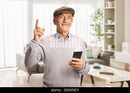Der ältere Mann zeigt nach oben und hält sein Smartphone zu Hause in einem Wohnzimmer Stockfoto