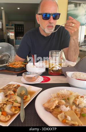 Ein Mann mittleren Alters, der eine Mahlzeit in einem asiatischen zwanglosen Restaurant genießt, umgeben von schmackhaften Gerichten und einem kalten Getränk Stockfoto
