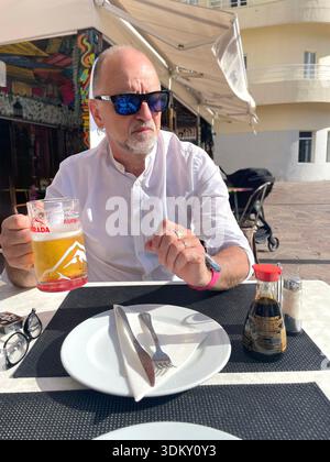 Ein Mann mittleren Alters sitzt an einem Café-Tisch im Freien mit einem Bier und sieht nachdenklich aus, während er ein Gericht in spanien genießt Stockfoto
