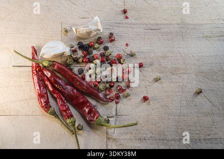Rote Chilischoten, Knoblauchzehen und eine Mischung aus schwarzen und grünen Pfefferkörnern sind auf einem Holzbrett angeordnet. Diese Zutaten sind gebrauchsfertig Stockfoto