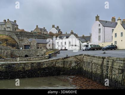 Alte Steinhäuser und weiß getünchte Häuser bilden eine malerische Straße in der Fischerstadt Portsoy. Die Autos parken am Shore Inn und die Hafenmauern treffen auf tr Stockfoto