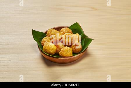 Goldene Kartoffelknusperbällchen serviert in einer rustikalen Holzschale, die mit frischen Bananenblättern auf hellem Holzhintergrund gesäumt ist Stockfoto