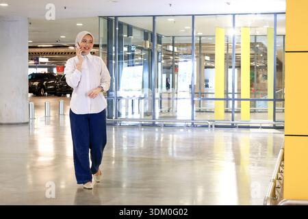 Junge Hijab-Frau, die auf dem Parkplatz läuft, während sie einen Anruf tätigt Stockfoto