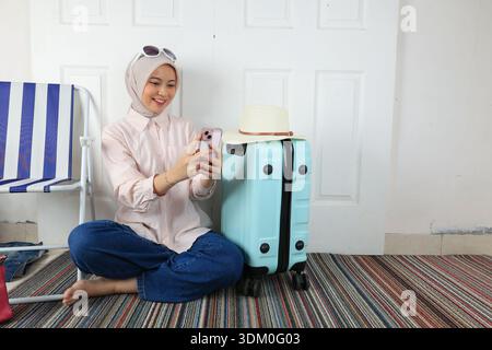 Die junge asiatische Hijab-Frau bereitet Reisesachen vor, überprüft die Checkliste auf ihrem Handy Stockfoto