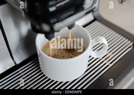 Professioneller Espresso, der in eine weiße Keramiktasse fließt, aus der modernen Kaffeemaschine im Catering-Buffet oder in der Büroküche Stockfoto