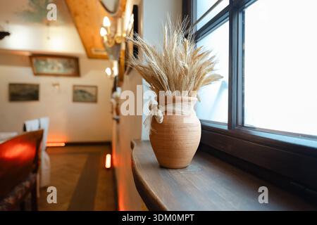 Rustikale Tonvase mit getrocknetem Weizen auf einer Fensterbank in gemütlicher Einrichtung mit warmem Licht und traditioneller Einrichtung Stockfoto