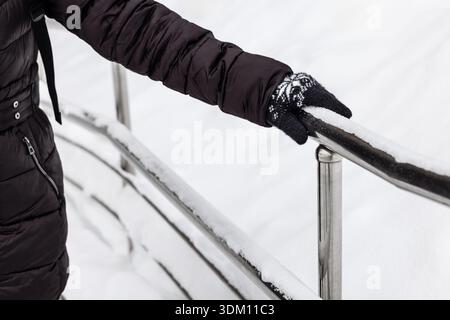 Eine Frau hält sich im Winter an einem Handlauf aus Metall. Eine Frau in Winterkleidung hält sich an einem mit Schnee bedeckten Handlauf fest. Geländer im Winterpark. Sicherheit in Stockfoto