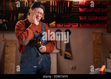 Kranke Tischlerin trägt Arbeitskleidung in der Werkstattszene mit Werkzeugen, die an der Wand hängen und Zahnschmerzen leiden. Zimmerfrau am späten Abend in authentischer Garage. Stockfoto