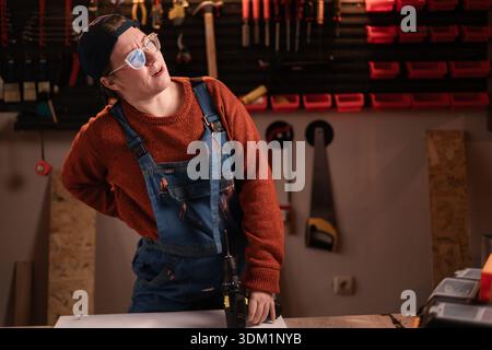 Gesundheitskonzept. Die Tischlerin trägt Arbeitskleidung in der Werkstattszene mit Werkzeugen, die an der Wand hängen und an akuten Rückenschmerzen leiden. Zimmerfrau spät Stockfoto
