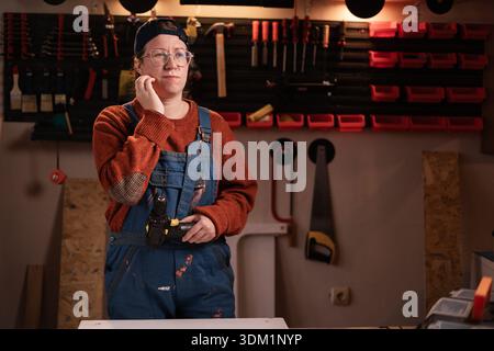 Kranke Tischlerin trägt Arbeitskleidung in der Werkstattszene mit Werkzeugen, die an der Wand hängen und Zahnschmerzen leiden. Zimmerfrau am späten Abend in authentischer Garage. Stockfoto
