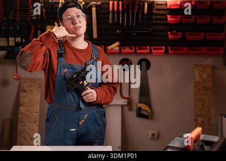Die Tischlerin trägt Arbeitskleidung in der Werkstattszene mit Werkzeugen, die an der Wand hängen, die eine Telefongeste zeigen, sagen Sie, ich rufe mich an. Zimmermanninnen am späten Abend in authentischer Atmosphäre Stockfoto