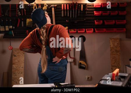 Gesundheitskonzept. Die Tischlerin trägt Arbeitskleidung in der Werkstattszene mit Werkzeugen, die an der Wand hängen und an akuten Rückenschmerzen leiden. Zimmerfrau spät Stockfoto
