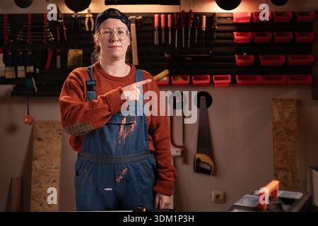Sehen Sie sich an. Tischlerin trägt Arbeitskleidung in der Werkstattszene mit Werkzeugen, die an der Wand hängen und mit dem Finger auf den Kopierraum zeigen. Zimmerfrau am späten Abend Stockfoto