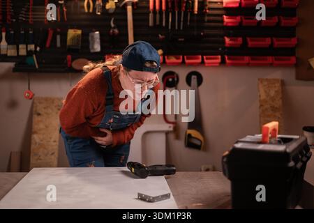 Gesundheitskonzept. Die Tischlerin trägt Arbeitskleidung in der Werkstattszene mit Werkzeugen, die an der Wand hängen, und leidet unter Magenschmerzen. Zimmerfrau spät ev Stockfoto