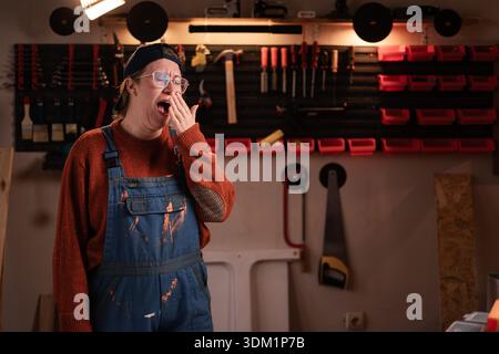 Die Tischlerin trägt Arbeitskleidung in der Werkstattszene mit Werkzeugen, die an der Wand hängen, während sie vor der Kamera posiert. Zimmerfrau am späten Abend in authentischer Ga Stockfoto