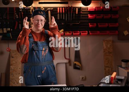 Die Tischlerin trägt Arbeitskleidung in der Werkstattszene, wobei die Werkzeuge an der Wand hängen, indem sie mit dem Finger gekreuzt wird. Zimmerfrau am späten Abend in authentischer Garage. Stockfoto