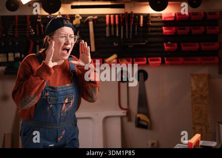 Die Tischlerin trägt Arbeitskleidung in der Werkstattszene mit Werkzeugen, die an der Wand hängen, schreiende Ansage. Zimmerfrau am späten Abend in authentischer Garage. Co Stockfoto
