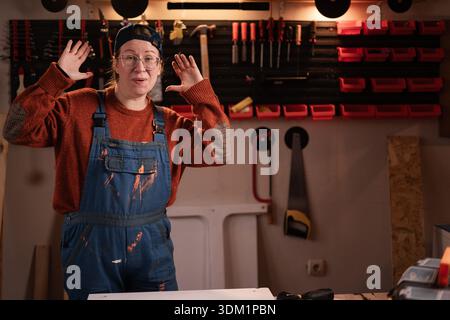 Glückliche Tischlerin trägt Arbeitskleidung in der Werkstattszene mit Werkzeugen, die an der Wand hängen, lächelnd in die Kamera schauen. Zimmermanninnen am späten Abend in authentischer Atmosphäre Stockfoto