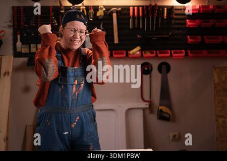 Die Tischlerin trägt Arbeitskleidung in der Werkstattszene mit Werkzeugen, die an der Wand hängen und die Siegergesten feiern. Zimmerfrau am späten Abend Stockfoto