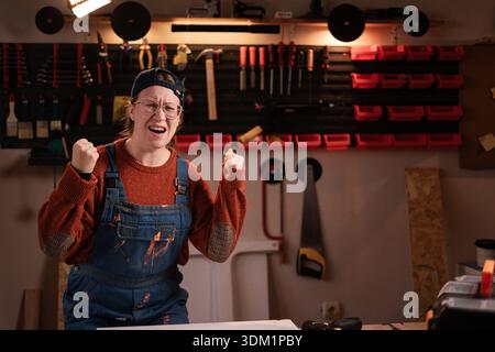 Die Tischlerin trägt Arbeitskleidung in der Werkstattszene mit Werkzeugen, die an der Wand hängen und die Siegergesten feiern. Zimmerfrau am späten Abend Stockfoto
