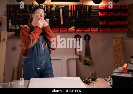 Schockiert staunte Tischlerin trägt Arbeitskleidung in der Werkstattszene mit Werkzeugen, die an der Wand hängen, hat einen Schock für Nachrichten. Zimmermanninnen am späten Abend in authentischer Atmosphäre Stockfoto