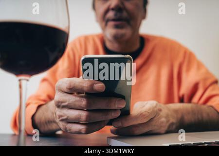 Ein Mann mittleren Alters in einem orangefarbenen Pullover sitzt mit Laptop und einem Glas Rotwein an einem Tisch und fokussiert sich auf seinen Smartphone-Bildschirm, um das moderne t zu veranschaulichen Stockfoto