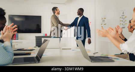 Zwei verschiedene männliche Führungskräfte schütteln in einem hellen Büro die Hand, während Kollegen applaudieren und für professionelle Anerkennung und integrative Führung stehen Stockfoto