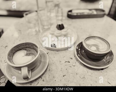 Zwei Kaffeetassen mit aufwändiger Latte Art stehen auf einem Marmortisch neben einem Dessertteller mit Gebäck und Gläsern Wasser in einem Café Stockfoto