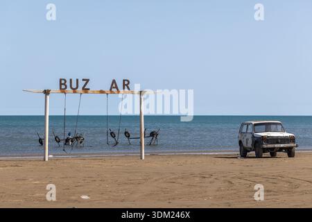 Buzowna, Aserbaidschan - 29. März 2025: Ein altes, verwittertes weißes Vintage-Auto aus dem Jahr 1987 von Lada Niva steht am Sandstrand nahe der rostigen Schaukel Stockfoto