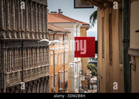 Leeres, rotes quadratisches Schild, das über einer schmalen Straße in einer Stadt im mediterranen Stil hängt und Kopierraum für Branding oder Werbung bietet, mit altem Gebäude Stockfoto