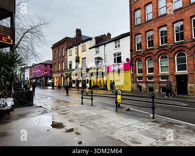 Manchester, Großbritannien - 21. Januar 2026: Bunte Ladenfronten säumen eine regnerische Manchester Street mit Fußgängern und urbanem Leben. Stockfoto
