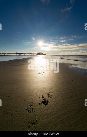 Sonnenaufgang über dem Worthing Pier vom Strand mit Fußabdrücken auf reflektierendem nassem Sand und strahlendem Horizont Stockfoto