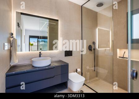 Stilvolles beigefarbenes Badezimmer mit Waschbecken, Duschkabine, schwarzen Armaturen und beleuchtetem Spiegel Stockfoto