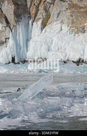 Scharfe türkisfarbene Eissplitter liegen auf der gefrorenen Oberfläche des Baikalsees mit massiven, eisbedeckten Klippen im Hintergrund unter klarem Winterlicht Stockfoto