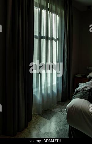 Sanftes Morgenlicht filtert durch weiße Vorhänge und dunkle Vorhänge in einem Hotelzimmer und wirft Schatten über einen Marmorboden in Valencia, Spanien. Stockfoto