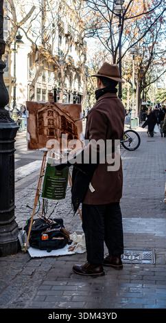 Der Straßenkünstler Sandro M. Miralles trägt einen Fedora und einen braunen Mantel, während er an seiner Staffelei auf einem Bürgersteig in Valencia, Spanien, ein monochromatisches Stadtbild malte. Stockfoto