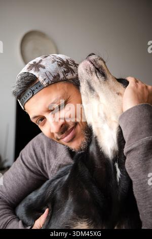 Ein Mann umarmt einen Hund mit einem schwarz-weiß gepunkteten Gesicht. Der Mann lächelt und der Hund wackelt mit dem Schwanz. Die Szene ist glücklich und liebevoll Stockfoto