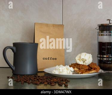 Ein warmes Stillleben in der Küche mit braunem Kaffeebeutel, dunklem Becher, verstreuten Bohnen, einem Teller mit Kuchen- und Vanilleeis, Schlagsahne und einem Glas Stockfoto