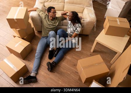 Ein junges schwarzes Paar drinnen mit Pappkartons, das sich über die Wohnungsarrangements unterhält. Freund und Freundin entspannen sich auf dem Boden, lehnen sich auf die Couch und genießen während des Umzugstages eine Pause. Stockfoto