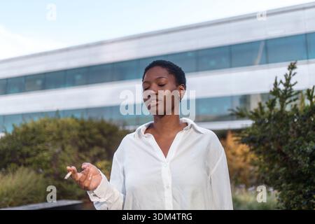 Junge Geschäftsfrau in weißem Hemd, die Zigarette hält, während sie vor einem modernen Bürogebäude steht und entspannt mit geschlossenen Augen aussieht Stockfoto