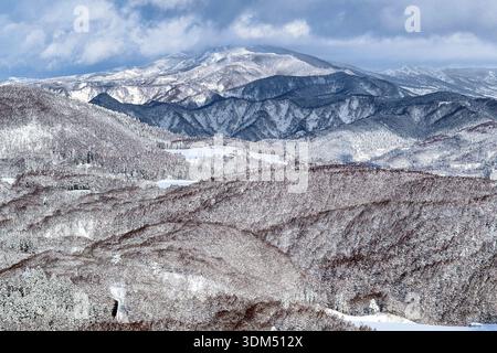 Panoramablick auf verschneite Hügel und ferne Berggipfel unter dramatisch bewölktem Winterhimmel Stockfoto