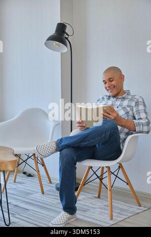 Ein lächelnder Mann, der sich in einem modernen Stuhl entspannt und mit seinem Buch eine ruhige Freizeit genießt Stockfoto