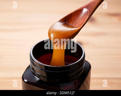 Dicker Manuka-Honig, der aus einem schwarzen Glas mit einem Holzlöffel auf einem Holztisch geschöpft wird. Stockfoto