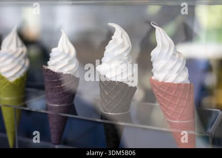 Verführerische Ausstellung von vier farbenfrohen, weichen Eiskegeln. Dieses köstliche Vanilledessert in grünen, lila und rosa Waffelkegeln wartet hinter Glas Stockfoto