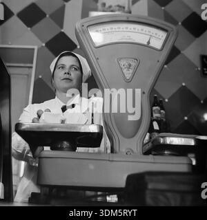 Slawjansk, Region Donezk, ukrainische SSR, UdSSR. 1970er Jahre Eine ausdrucksstarke Archivansicht einer Ladenassistentin, die ein Produkt in einem großen mechanischen Maßstab in einem Supermarkt wiegt. Die Frau in einem weißen Uniformmantel und einer Kappe legt ein gewickeltes Stück Butter oder Käse auf die Metallschale. Das Zifferblatt der schweren Industriewaage ist im Vordergrund deutlich zu erkennen. Das Bild fängt die Technologie des sowjetischen Handels, den Prozess des Kundenservice und die Atmosphäre einer Gastronomie-Abteilung im friedlichen Donbass ein. Stockfoto