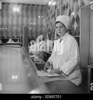 Slawjansk, Region Donezk, ukrainische SSR, UdSSR. 1970er Jahre Eine stimmungsvolle Archivinnenansicht der Gastronomie in einem städtischen Lebensmittelgeschäft. Eine Ladenassistentin in einem weißen Uniformmantel und einem hohen, stärkeren Hut steht hinter der Theke mit einem Messer und einem Geschenkpapier. Sie ist bereit, Butter oder Käse für Kunden zu schneiden. Im Hintergrund sind die Regale mit Pyramiden aus Konserven gefüllt. Eine weitere Verkäuferin arbeitet in der Nähe. Das Bild fängt die Ästhetik des sowjetischen Einzelhandels, die Fülle an Waren und die Servicekultur im friedlichen Donbass ein. Stockfoto