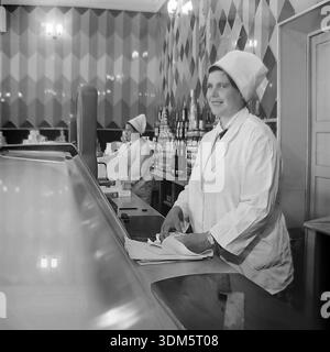 Slawjansk, Region Donezk, ukrainische SSR, UdSSR. 1970er Jahre Eine stimmungsvolle Archivinnenansicht der Gastronomie in einem städtischen Lebensmittelgeschäft. Eine Ladenassistentin in einem weißen Uniformmantel und einem hohen, stärkeren Hut steht hinter der Theke mit einem Messer und einem Geschenkpapier. Sie ist bereit, Butter oder Käse für Kunden zu schneiden. Im Hintergrund sind die Regale mit Pyramiden aus Konserven gefüllt. Eine weitere Verkäuferin arbeitet in der Nähe. Das Bild fängt die Ästhetik des sowjetischen Einzelhandels, die Fülle an Waren und die Servicekultur im friedlichen Donbass ein. Stockfoto