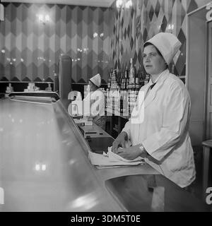 Slawjansk, Region Donezk, ukrainische SSR, UdSSR. 1970er Jahre Eine stimmungsvolle Archivinnenansicht der Gastronomie in einem städtischen Lebensmittelgeschäft. Eine Ladenassistentin in einem weißen Uniformmantel und einem hohen, stärkeren Hut steht hinter der Theke mit einem Messer und einem Geschenkpapier. Sie ist bereit, Butter oder Käse für Kunden zu schneiden. Im Hintergrund sind die Regale mit Pyramiden aus Konserven gefüllt. Eine weitere Verkäuferin arbeitet in der Nähe. Das Bild fängt die Ästhetik des sowjetischen Einzelhandels, die Fülle an Waren und die Servicekultur im friedlichen Donbass ein. Stockfoto
